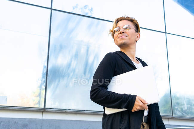 Снизу студент-мужчина в повседневной одежде и очках с современным нетбуком в руке, идущий по улице рядом с современным зданием — стоковое фото