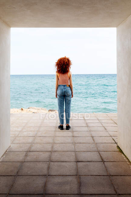 Обратный вид на всю длину неузнаваемой женщины-туристки, любующейся волнистым морем с бетонного прохода на побережье — стоковое фото