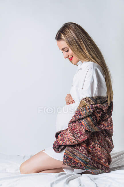 Боковой вид молодой нежной беременной женщины, касающейся живота, сидящей на кровати и счастливо улыбающейся — стоковое фото