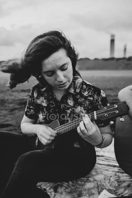 Талантливая женщина-музыкант с каштановыми волосами в повседневной одежде, играющая на укулеле и поющая песню, сидя днем на песчаном пляже на природе — стоковое фото