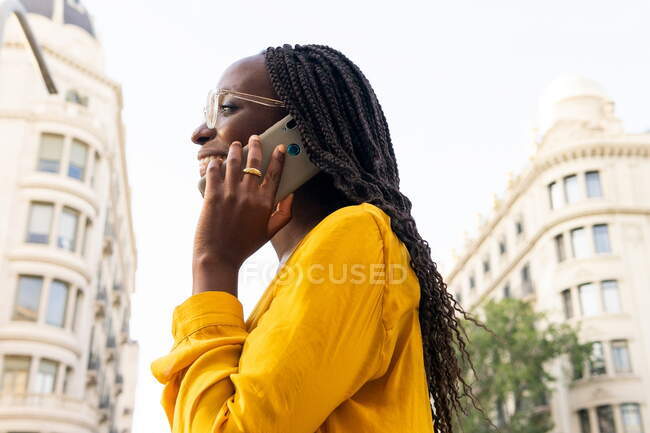Позитивна афроамериканка в окулярах, що відвернулася під час телефонної розмови на вулиці з житловими будинками і деревами в місті. — стокове фото