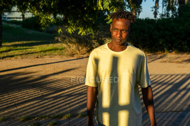 Jovem afro-americano masculino em roupas casuais em pé no beco no parque verde e olhando para longe pensativo ao pôr do sol luz — Fotografia de Stock