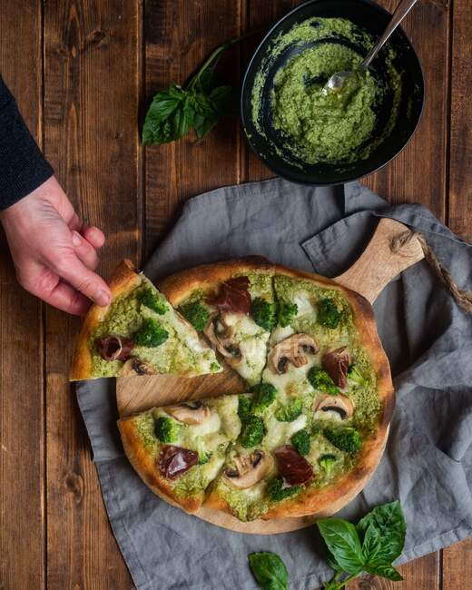 Top view of crop unrecognizable person taking slice of tasty pizza with pesto sauce and mushrooms served on wooden cutting board on table — Stock Photo