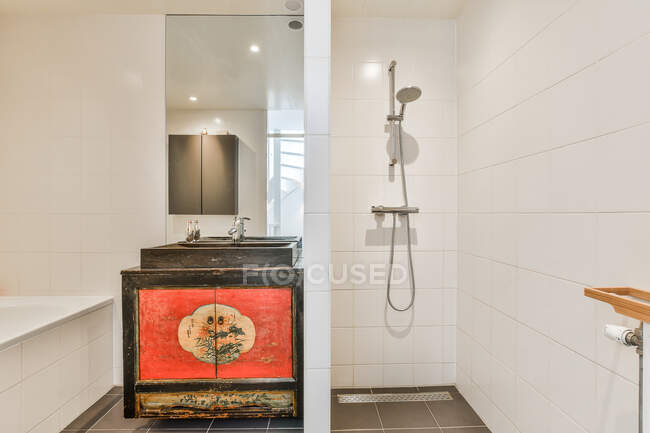 Ванна біля шафи з раковиною біля плиткової стіни з дзеркалом з відображенням у світлій стильній ванній кімнаті з душем і перегородкою — стокове фото