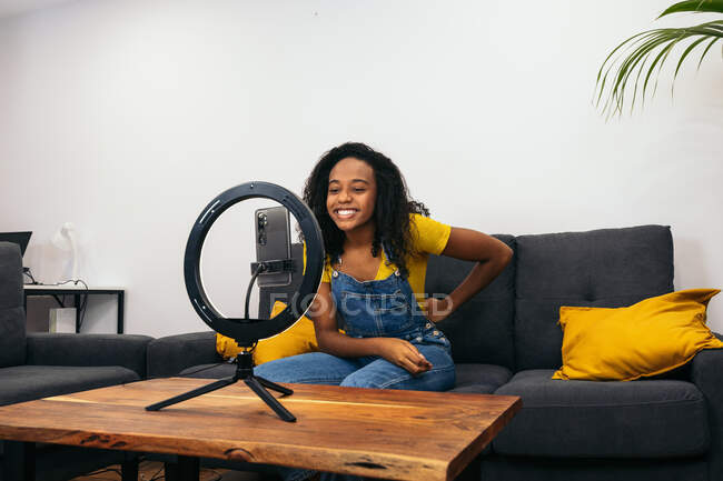 Sonriente hembra negra en el sofá mientras usa el teléfono inteligente en la lámpara de anillo led cerca de luces profesionales en trípodes - foto de stock