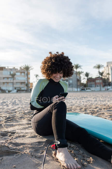 Femme positive avec les cheveux bouclés portant une combinaison de navigation smartphone tout en étant assis sur une plage de sable avec planche de surf — Photo de stock