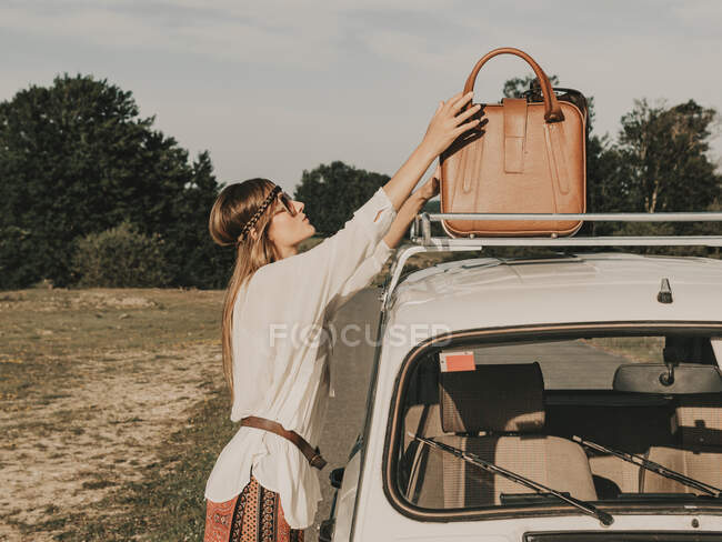 Side view of female hippie in sunglasses taking luggage from white old timer automobile parked in sunny nature during trip — Stock Photo