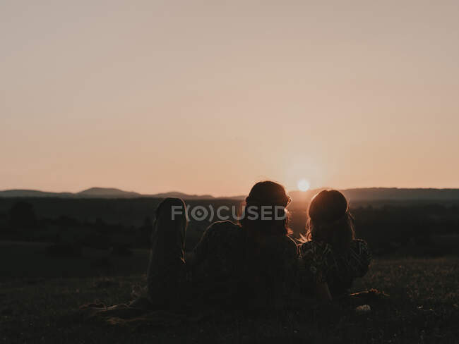 Пара хіпі з сонцезахисними окулярами, що лежать на трав'янистих лугах в природі, спостерігаючи за заходом сонця серед гір — стокове фото