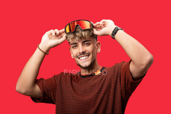 Позитивный молодой человек с вьющимися волосами в модных солнцезащитных очках широко улыбается на красном фоне и смотрит в камеру — стоковое фото
