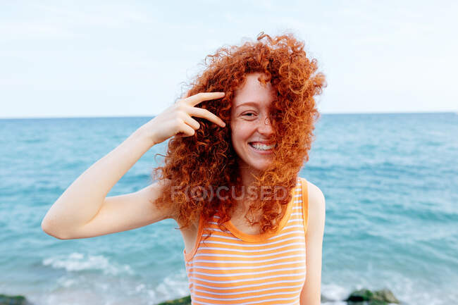 Mulher jovem otimista com cabelo de gengibre voador olhando para a câmera na costa do mar ondulante azul — Fotografia de Stock