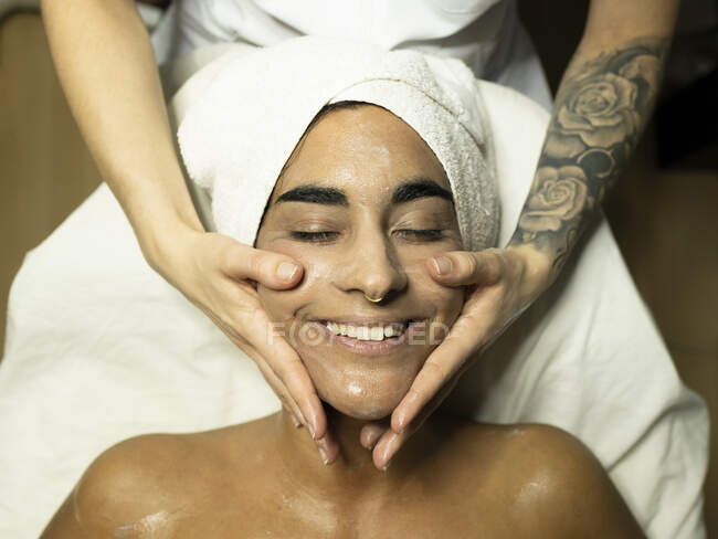 Женщина-массажистка наносит крем на лицо клиента, делая массаж лица на диване в ярком спа-салоне — стоковое фото