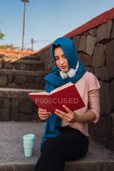 Hembra musulmana pensativa en hijab sentada en escaleras de piedra en el parque de la ciudad y leyendo un libro interesante durante el fin de semana - foto de stock