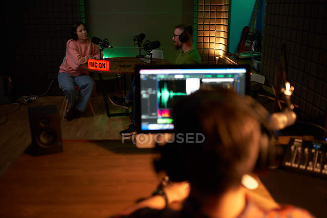 Back view of unrecognizable male sound director sitting at table with computer during podcast recording with host and female singer in dark studio — Stock Photo