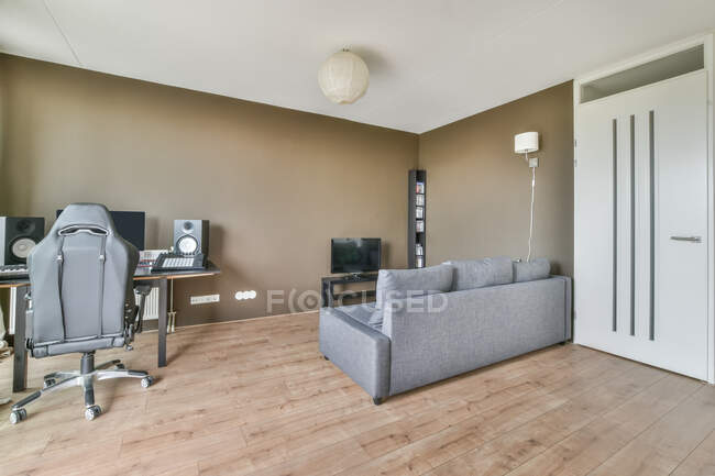 Interior de la moderna sala de estar con paredes de color marrón claro y suelo y sofá gris y TV y zona de trabajo con computadora de escritorio y altavoces y escritorio y sillón - foto de stock