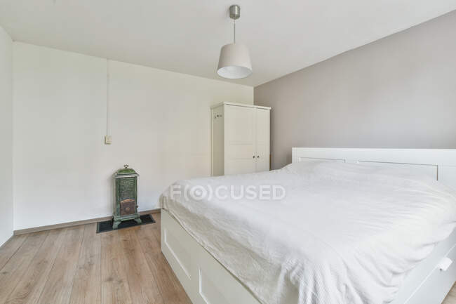 Interior del dormitorio amueblado con cama cubierta con cobertor blanco colocado cerca de armario de madera debajo de la lámpara - foto de stock