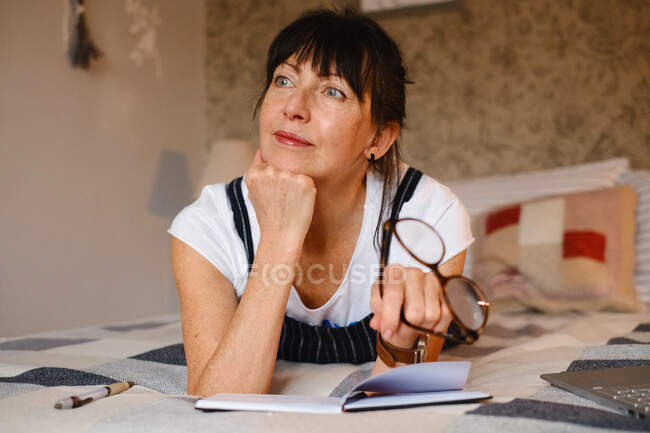 Femme d'âge moyen positive penchée sur la main alors qu'elle était couchée sur le lit avec un bloc-notes et des lunettes ouverts et regardant ailleurs — Photo de stock