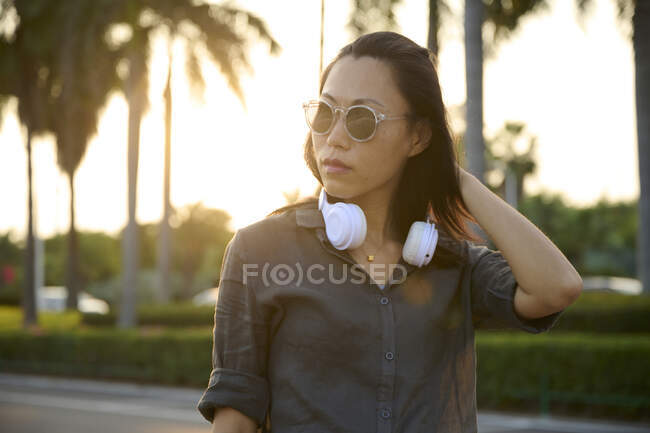 Serious Asian female with modern white headphones looking into distance while standing near road on street of town with green trees — Stock Photo