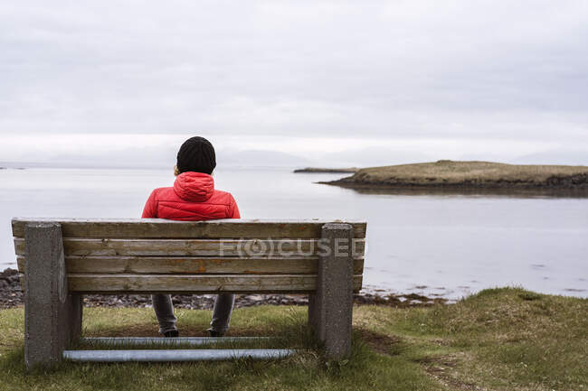 Vista trasera de una mujer irreconocible en ropa interior cálida sentada en un banco de madera en la orilla y disfrutando de pintorescos paisajes del océano bajo el cielo nublado en Islandia - foto de stock
