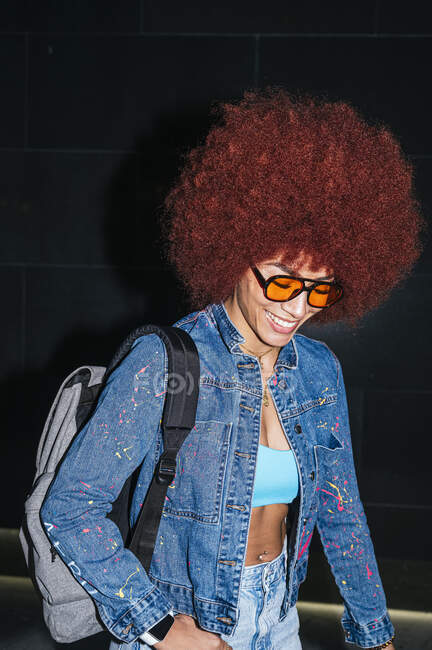 Lächelnde Frau mit Afro-Frisur und trendigem Outfit und Sonnenbrille, die den Boden betrachtet, während sie abends mit Rucksack auf schwarzem Hintergrund steht — Stockfoto