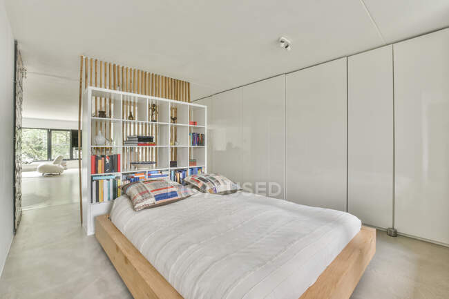 Bett mit Kissen auf dem Bezug gegen Regale mit Büchern und Dekor unter der weißen Decke zu Hause tagsüber — Stockfoto