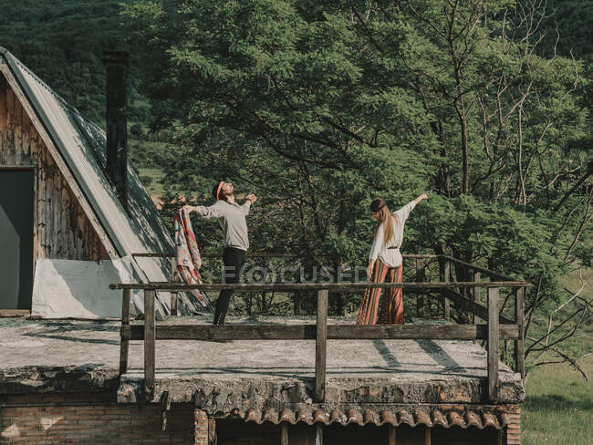 Full body of hippies in boho styled clothes dancing while standing on terrace near wooden cabin during trip in nature — Stock Photo