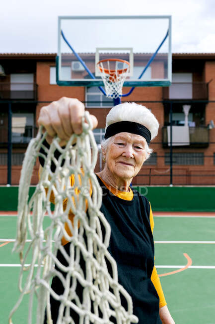 Senior hembra en ropa deportiva y diadema mirando a la cámara mientras está de pie con la red en la mano en la cancha de baloncesto con aro - foto de stock