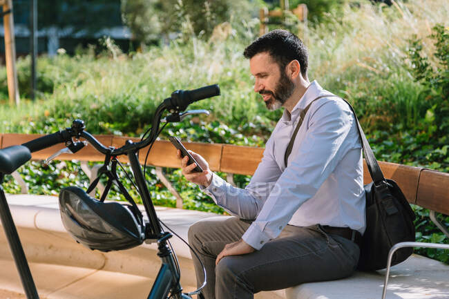 Side view of male manager sitting on bench near bicycle and reading messages on smartphone in park — Stock Photo