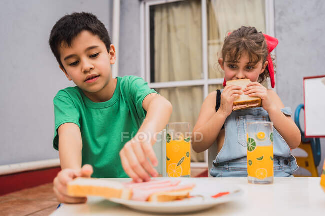 Bambini positivi in abbigliamento casual con panini freschi in mano seduti su sedie vicino a bicchieri nella stanza della luce a casa — Foto stock