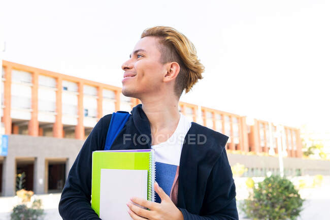 Side view of male student with workbooks looking away with thoughtful face while standing near modern university building on street during studies — Stock Photo
