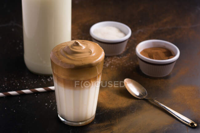Ein Glas köstlichen Dalgona-Kaffees mit Milch und schaumigem Belag auf einem schwarzen Tisch mit Kakaopulver und Zucker — Stockfoto