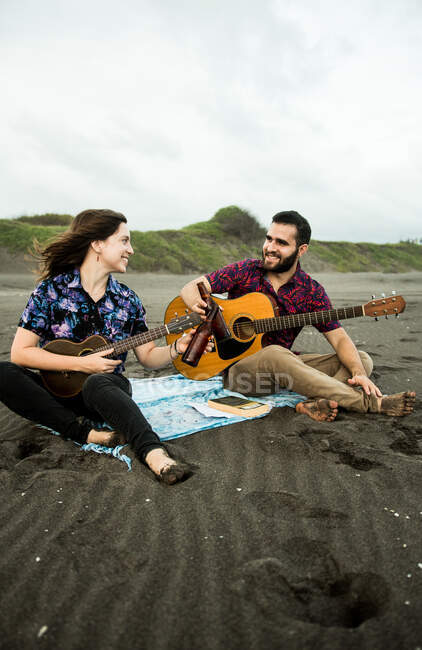 Позитивная пара музыкантов, сидящих с гитарами и звенящими бутылками пива, сидя днем на песчаном пляже возле океана — стоковое фото
