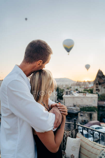 Side view of loving man embracing woman from behind looking away on roof terrace with hot air balloons in evening sky in Cappadocia Turkey — Stock Photo