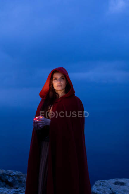 Serious female in Victorian styled outfit with red cape looking away while standing with glowing candle in hands in dark nature — Stock Photo