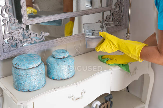 Crop unrecognizable female housekeeper in apron and rubber gloves wiping decorative items placed on cabinet under mirror in hall during work in elegant apartment — Stock Photo