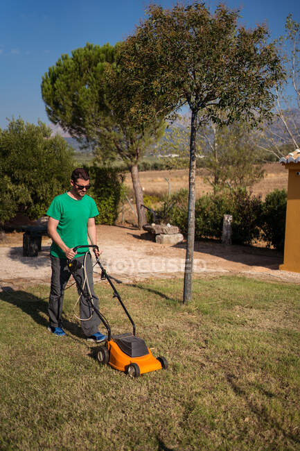 Visão lateral de corpo inteiro do jardineiro masculino no chapéu cortando gramado perto de arbustos e árvores no verão — Fotografia de Stock