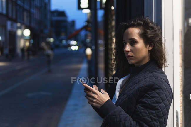 Side view of thoughtful female in warm clothes using smartphone while standing on street and looking away in evening — Stock Photo