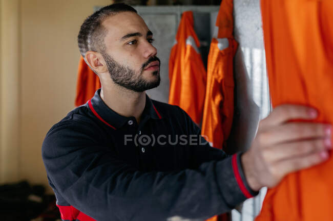 Молодой красивый бородатый этнический мужчина открывает шкафчик и готовится надеть оранжевую форму на пожарную станцию — стоковое фото