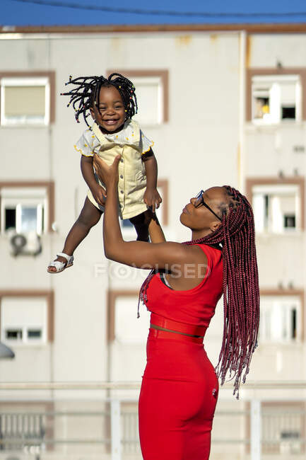 Side view of African American female with long braids in stylish outfit standing and lifting happy little daughter while having fun on street in sunny day — Stock Photo