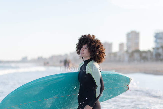 Боковой вид молодой вдумчивой серфингистки в гидрокостюме с доской для сёрфинга, стоящей на берегу моря, умытой морем — стоковое фото