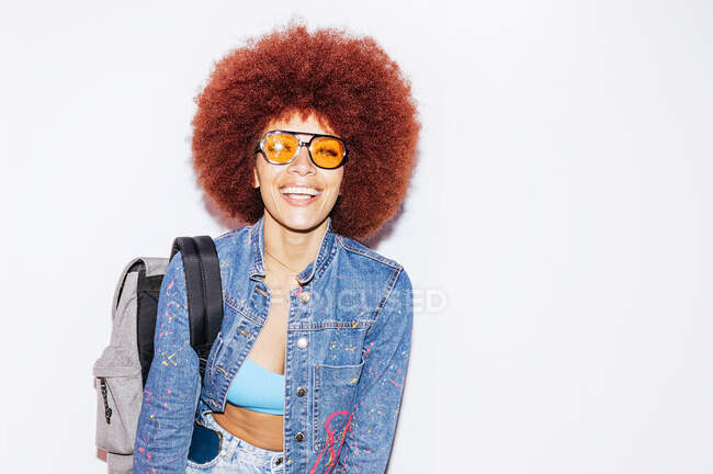 Positive female with modern hairstyle wearing trendy outfit and sunglasses looking at camera while standing on white background with backpack — Stock Photo
