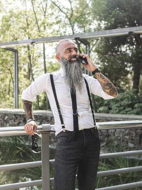 Confident smiling bearded hipster in stylish outfit speaking on cellphone while standing near metal fence on sunny street — Stock Photo
