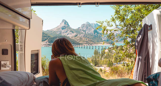 Back view of unrecognizable female traveler lying on bed in van and enjoying scenery of blue lake located in highlands in Riano — Stock Photo