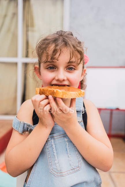 Carino ragazza in denim generale guardando la fotocamera mentre si mangia pane fresco con marmellata dolce nella stanza luce a casa — Foto stock