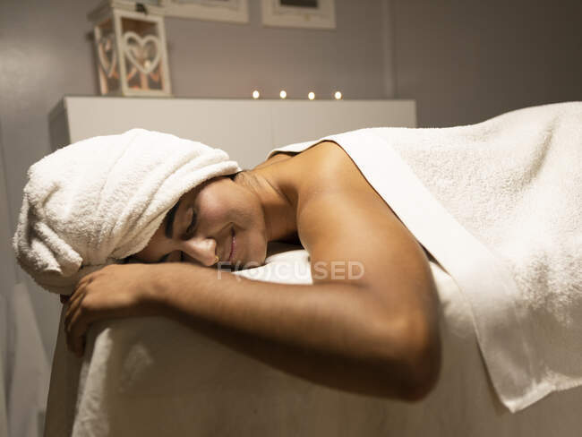 Positivo giovane signora etnica sotto asciugamano sdraiato sul divano con gli occhi chiusi nel salone leggero spa — Foto stock