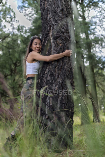 Спокойная женщина в повседневной одежде с закрытыми глазами, обнимающая толстый ствол дерева в лесу с размытой зеленой травой во время похода — стоковое фото