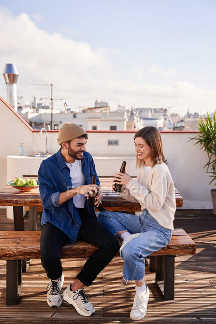 Positivos jovens companheiros de apartamento masculinos e femininos sentados no banco com garrafas de cerveja e tendo uma conversa agradável no terraço — Fotografia de Stock