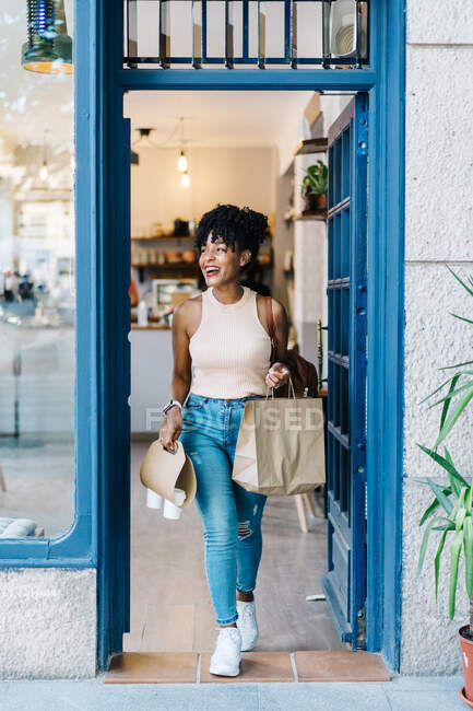 Ganzkörper positive junge Afroamerikanerin mit lockigem Haar in lässiger Kleidung lächelt, während sie ein modernes Café mit Kaffeetassen zum Mitnehmen und Papiertüten verlässt — Stockfoto