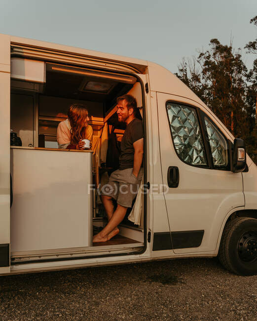 Cheerful couple of travelers standing in parked camper and looking at each other while enjoying sunset in summer during road trip in Galicia — Stock Photo