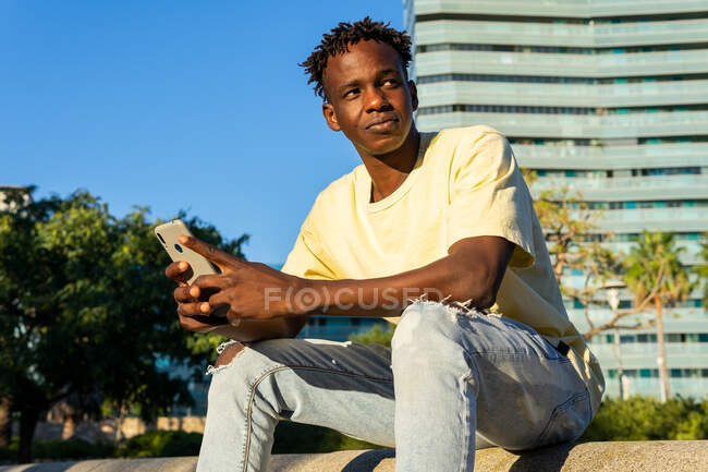 Thoughtful African American male wearing casual clothes sitting on street barrier while chatting on smartphone — Stock Photo