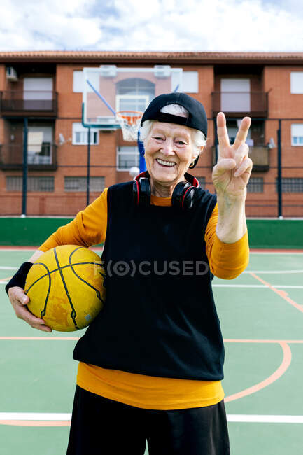 Оптимистичная зрелая женщина в спортивной одежде и наушниках смотрит в камеру, стоя на общественной баскетбольной площадке с мячом во время тренировки — стоковое фото